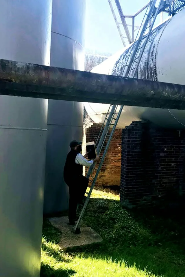 Representative on a ladder beneath vertical storage tanks, reviewing documents during a site inspection